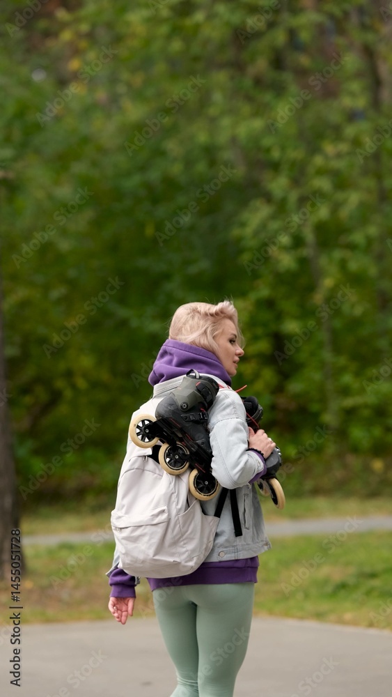 Fototapeta premium Beautiful blonde woman walking through city park holding roller skates enjoying beautiful day
