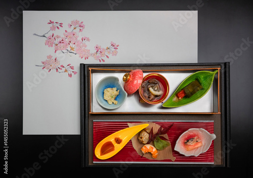A beautifully arranged Japanese kaiseki meal is presented on a black tray with a cherry blossom-themed placemat, captured from a top-down perspective to emphasize the elegance and detail of each dish.