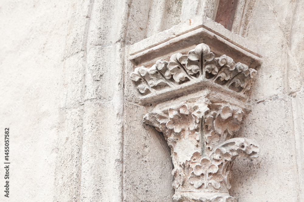Intricately carved stone capital adorns top of column on historic building, featuring floral and leaf motifs. Weathered texture of stone contrasts with smooth wall, highlighting aged craftsmanship