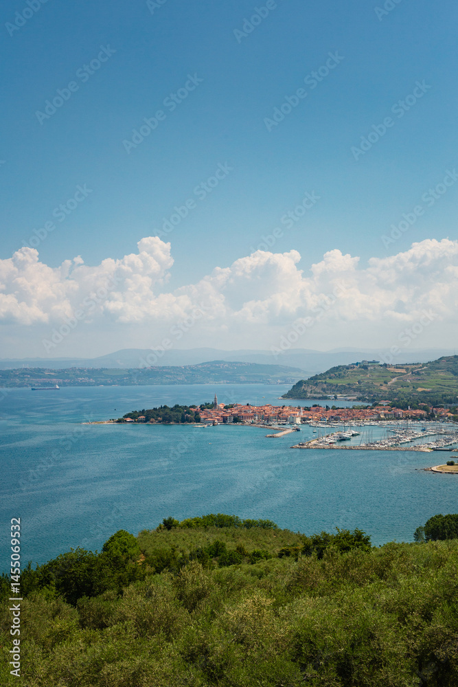Naklejka premium A scenic panoramic view of the coastal town of Izola in Slovenian Istria, featuring the harbor, red-tiled roofs, and the blue Adriatic Sea under a partly cloudy sky.