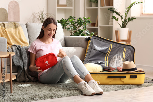 Young happy woman with suitcase and first aid kit sitting on floor at home. T...