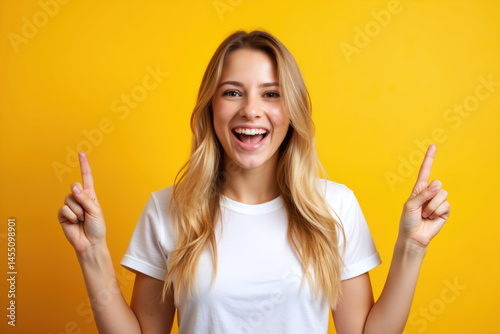Obraz na płótnie A cheerful woman with blonde hair, smiling broadly while pointing upwards with both hands against a bright yellow background. w salonie