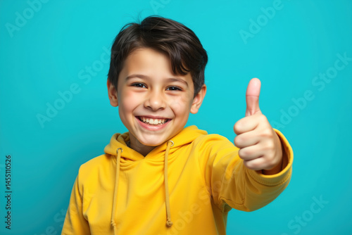 Obraz na płótnie A cheerful young boy wearing a yellow hoodie, smiling brightly, and giving a thumbs up against a vibrant blue background. w salonie