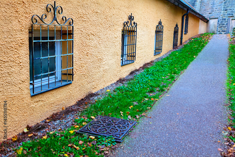 Narrow paved path runs alongside a yellow building with small windows framed by ornate black iron grilles. Path is bordered by green grass and decorative drain cover, with fallen autumn leaves