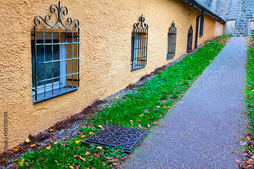Obraz na płótnie Narrow paved path runs alongside a yellow building with small windows framed by ornate black iron grilles. Path is bordered by green grass and decorative drain cover, with fallen autumn leaves w salonie