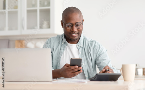 Savings and finances concept. Mature black man using calculator, phone and laptop computer, calculating taxes, sitting in kitchen at home, copy space