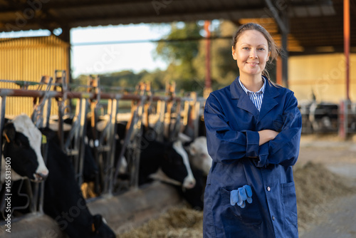 Portrait of woman ranch worker standing in cowshed near rows of cows in stalls with arms crossed.