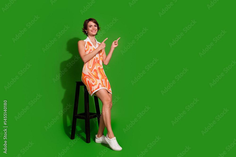 Full body photo of lovely young lady sit chair point empty space dressed stylish colorful garment isolated on green color background Full body photo of lovely young lady sit chair point empty space dressed stylish colorful garment isolated on green color background