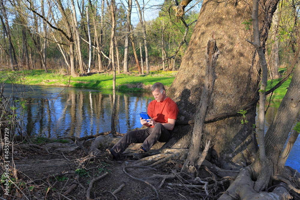 Middle-aged man in a red shirt finds a peaceful place by a stream to read Middle-aged man in a red shirt finds a peaceful place by a stream to read