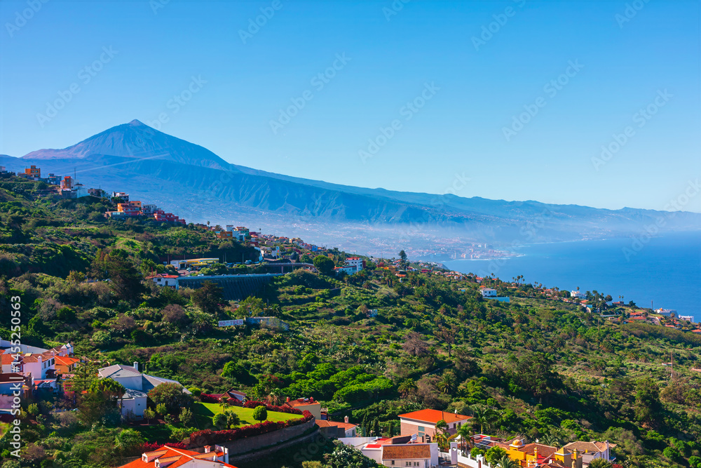 Lush green hills dotted with colorful houses cascade toward sparkling Atlantic with majestic Mount Teide rising in background. Clear sky and vivid contrast highlight Tenerife natural beauty
