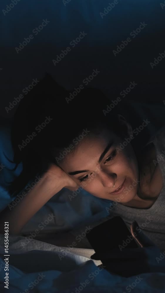 Vertical video. Mobile night. Gadget insomnia. Relaxed smiling woman enjoying using phone app online scrolling feed in bed late in dark home bedroom. Vertical video. Mobile night. Gadget insomnia. Relaxed smiling woman enjoying using phone app online scrolling feed in bed late in dark home bedroom.