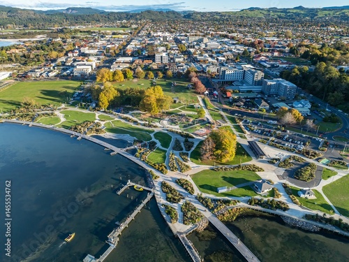 Aerial view of Rotorua, New Zealand, showcasing the city's urban landscape and the scenic Lake Rotorua. People enjoy the park, highlighting the city's blend of nature and urban life.
