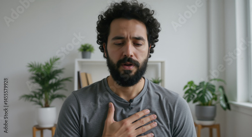 Man with curly hair and beard placing hand on chest with emotional expression in bright home setting. Heartfelt sincerity for personal testimony