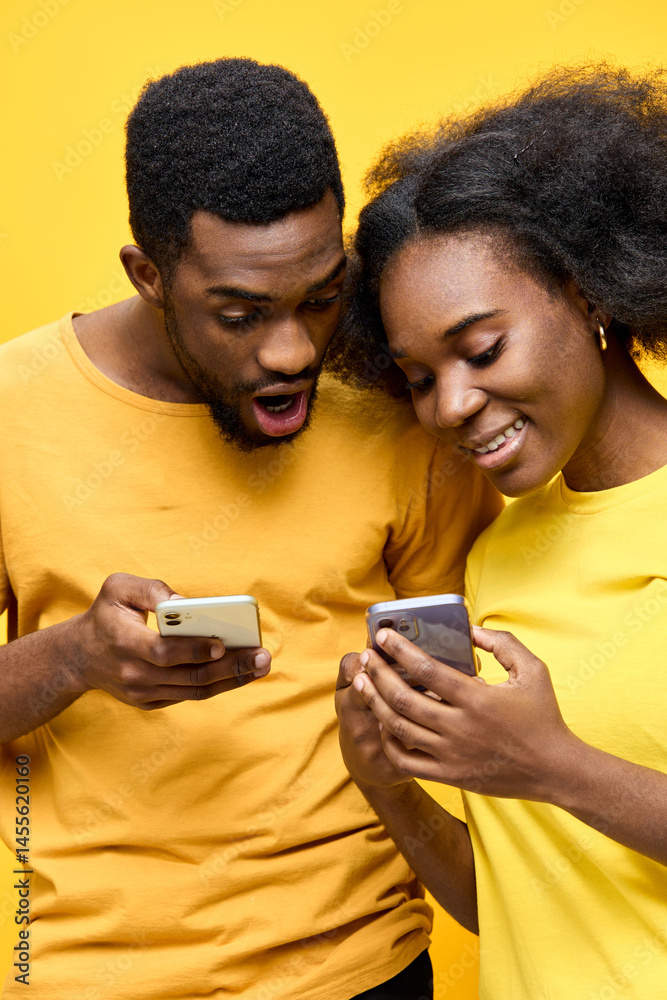 Fototapeta premium Young African couple joyfully interacting with smartphones against a bright yellow background, showcasing excitement and modern lifestyle choices