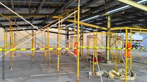 A construction site with yellow scaffolding under a metallic roof, surrounded by safety tape. 