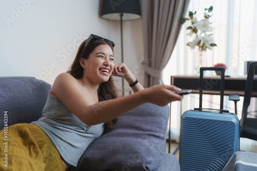 Young woman tourist is lying on the sofa in a hotel room, holding remote control and laughing while watching television after arriving from trip with her luggage
