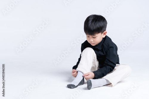 Young boy sitting on the floor and putting on socks, dressed in a black shirt and light pants, in a bright white studio background.