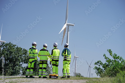 Maintenance engineer team standing at windmills at wind turbine farm. Group of people wear safety helmet and uniform working at alternative renewable energy wind station. Sustainable energy technology
