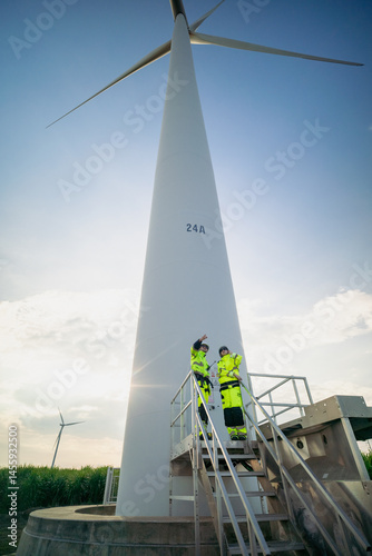 Maintenance engineer team standing at windmills at wind turbine farm. Group of people wear safety helmet and uniform working at alternative renewable energy wind station. Sustainable energy technology