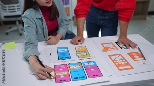 Two ux designers are analyzing paper prototypes of a mobile application interface, testing user flows and improving design elements on a white table