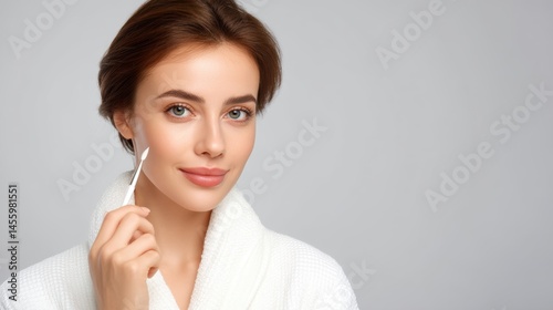 Obraz na płótnie A woman in a white robe applies skincare cream to her face with a spatula, smiling softly against a light background. w salonie