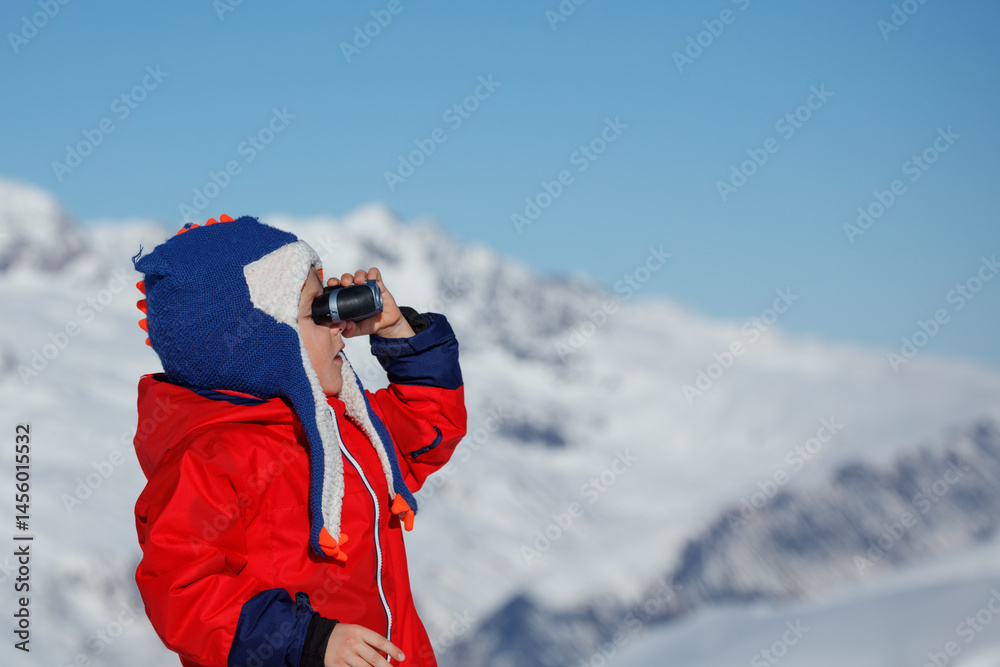 Young kid boy in blue funny hat views snowy peaks at binocular