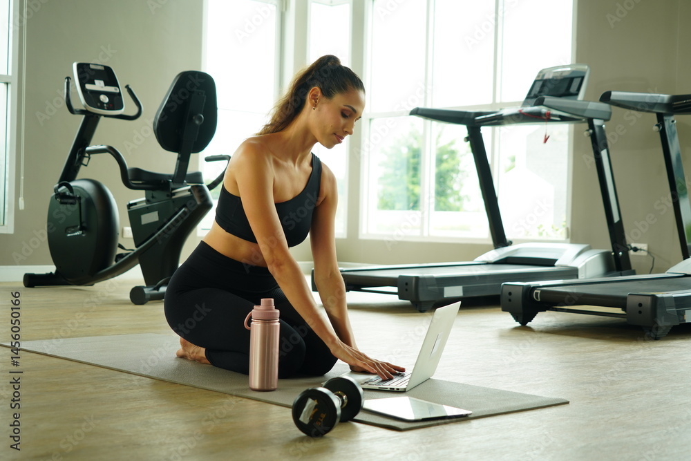 Beautiful Latin woman with a good figure sitting in a gym looking at a laptop to practice yoga exercise for health and weight loss in a fitness center.