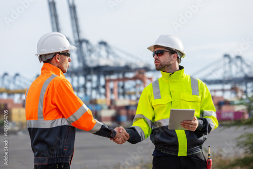 Two port authority officers wearing safety gear and helmets shake hands at a busy shipping port, signifying a successful agreement or partnership amid cranes and stacked cargo containers.