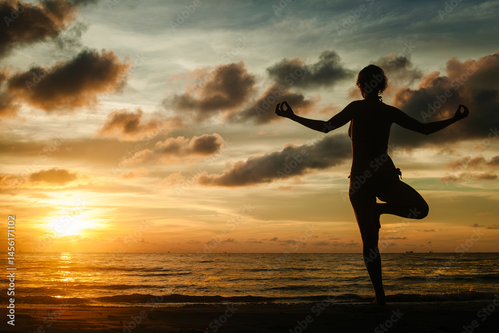A woman practicing yoga while standing on a tropical beach during a stunning sunset, surrounded by serene beauty and warm light.