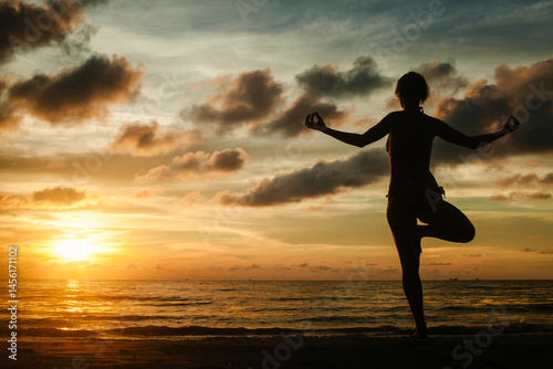 Obraz na płótnie A woman practicing yoga while standing on a tropical beach during a stunning sunset, surrounded by serene beauty and warm light. w salonie