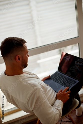 A man is working on a laptop by a large window. Man sitting in chair with laptop and smiling while working by a large window on a sunny day.