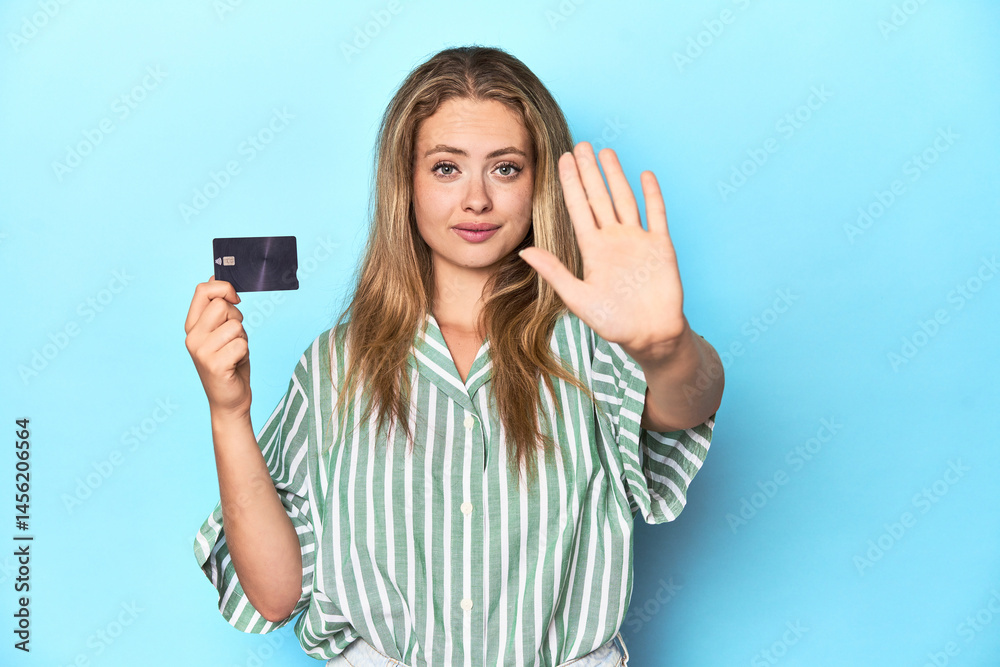 Young blonde with a credit card in blue studio standing with outstretched hand showing stop sign, preventing you. Young blonde with a credit card in blue studio standing with outstretched hand showing stop sign, preventing you.