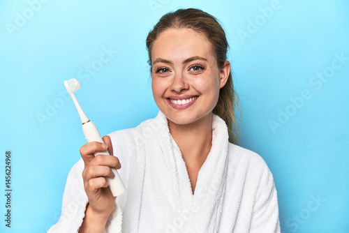 Young blonde woman in a robe holding an electric toothbrush in a studio