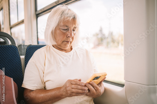 Obraz na płótnie A senior woman wearing a pink hat is sitting on a bus, using her smartphone. She is traveling through the city, enjoying her journey in a relaxed manner. w salonie