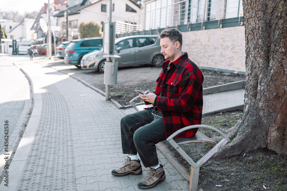 Male student with smartphone wearing plaid shirt sitting on bench while waiting for transport at tram or bus station. City public transport infrastructure, citizen and tourists transportation concept