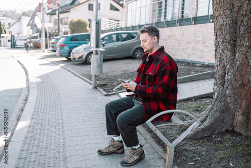 Obraz na płótnie Male student with smartphone wearing plaid shirt sitting on bench while waiting for transport at tram or bus station. City public transport infrastructure, citizen and tourists transportation concept w salonie