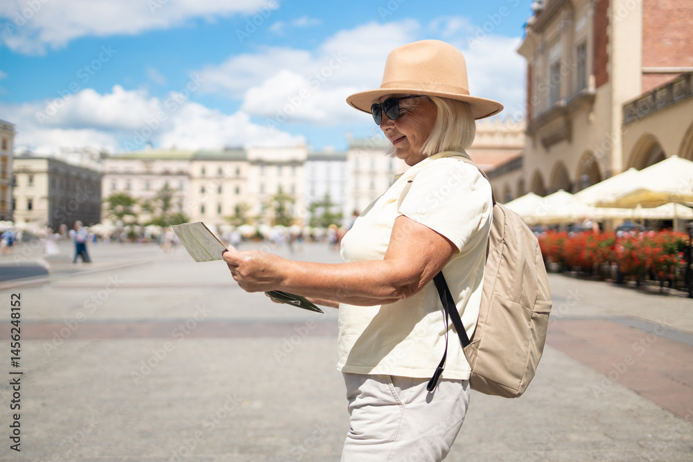 A senior woman stands in a beautiful city square, holding a map while enjoying sightseeing. She is dressed in a comfortable outfit, wearing a hat and sunglasses.