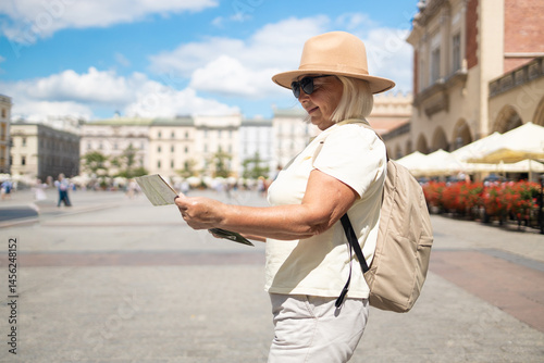 Obraz na płótnie A senior woman stands in a beautiful city square, holding a map while enjoying sightseeing. She is dressed in a comfortable outfit, wearing a hat and sunglasses. w salonie
