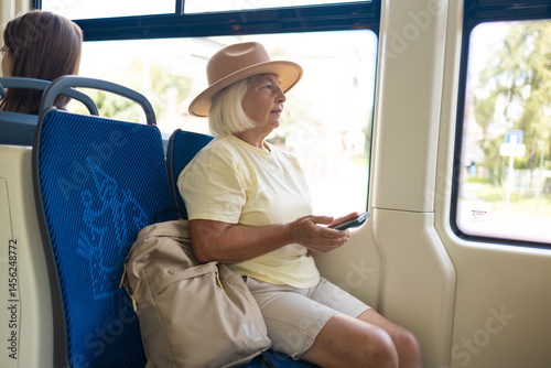 Obraz na płótnie Senior woman with a hat and a backpack is sitting in a tram, holding a smartphone and looking at the screen. w salonie