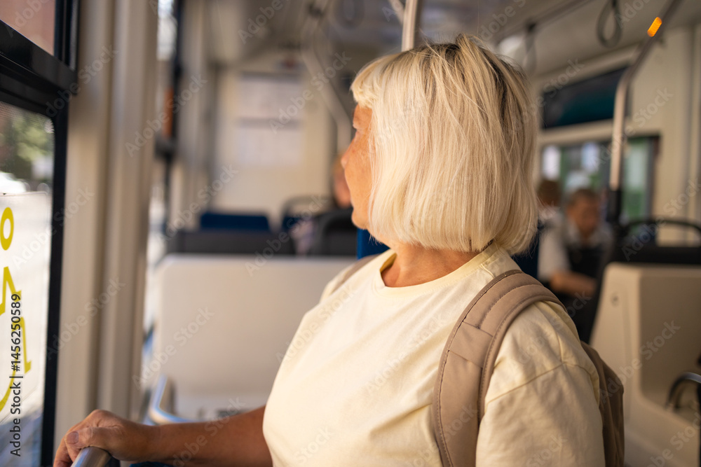 A senior woman with a backpack is standing by the window inside a tram, enjoying the view while traveling. She smiles as she looks out of the window.