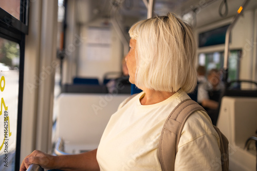 Obraz na płótnie A senior woman with a backpack is standing by the window inside a tram, enjoying the view while traveling. She smiles as she looks out of the window. w salonie