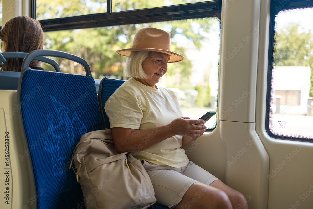 A senior woman wearing a pink hat is sitting on a bus, using her smartphone. She is traveling through the city, enjoying her journey in a relaxed manner.