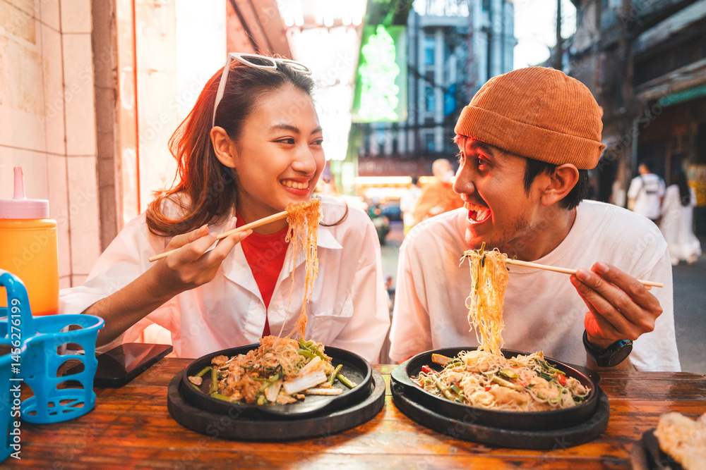 Obraz premium Asian couple man and woman enjoy eating street food noodle at night market. Traveler Asian couple blogger Happy tourists with Traditional food local road at thailand bangkok city.