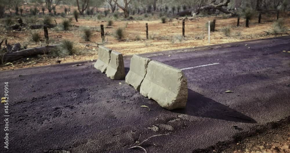 A weathered road barrier sits on a paved road, surrounded by dry vegetation and sparse trees in a remote outback location, indicating a potentially abandoned area.