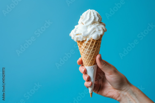 Obraz na płótnie A hand holding a vanilla ice cream cone against a bright blue background, showcasing a delicious summer treat. w salonie