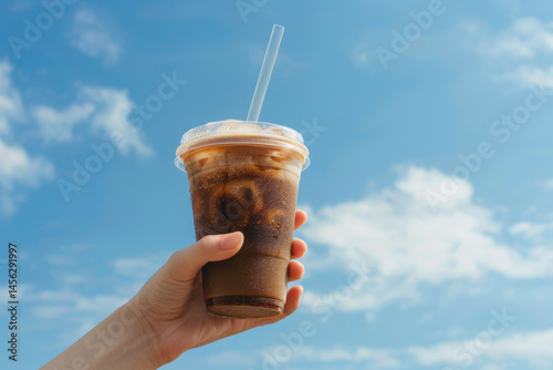 Obraz na płótnie A hand holding a cold iced coffee in a plastic cup with a straw against a bright blue sky. w salonie