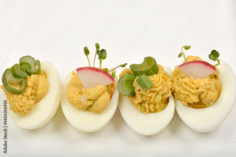 Four deviled egg halves, each uniquely garnished with celery, radish, and sprouts, are arranged on a white surface
