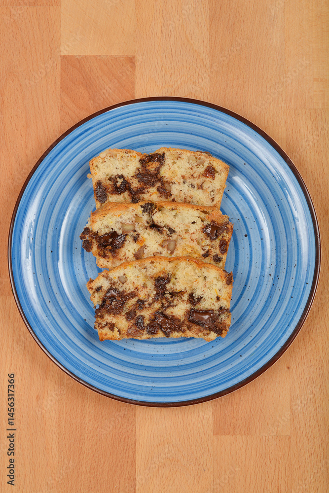 Three slices of a light-colored cake with visible pieces of chocolate, walnuts, raisins, and orange peel are arranged on a blue plate over a wooden surface