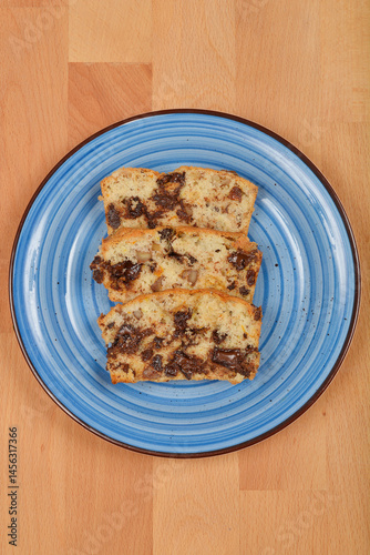Obraz na płótnie Three slices of a light-colored cake with visible pieces of chocolate, walnuts, raisins, and orange peel are arranged on a blue plate over a wooden surface w salonie