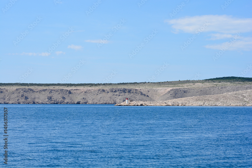 A view from Jadranovo shows Cape Bejavec and Turnac Lighthouse on Krk Island under a blue sky with the Adriatic Sea in the foreground